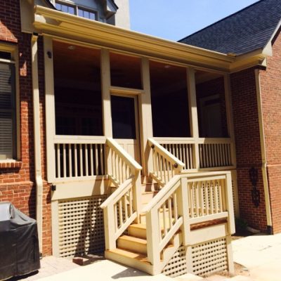 A brick house with a small porch featuring six steps, wooden railings, and a Painting Plus covered entrance. A grill is placed on a paved area to the left.