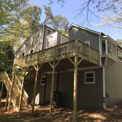 The Painting Plus two-story gray house features a charming wooden deck and staircase at the back, all beautifully surrounded by trees.