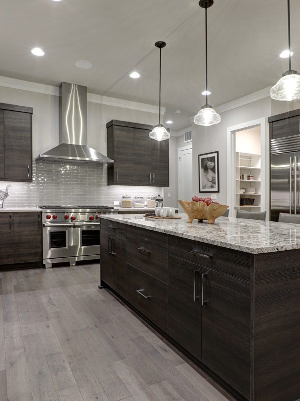 Modern kitchen with dark wood cabinets, stainless steel appliances, a large granite island, pendant lighting, light wood flooring, and interior paint colors that enhance the sleek design.